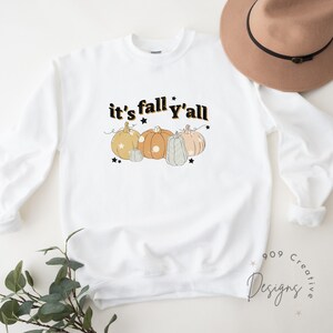 Puede incluir: Sudadera blanca con el texto "it's fall y'all" en negro con una fuente retro. El texto est&aacute; rodeado de cuatro calabazas en varios tonos de naranja y marr&oacute;n con estrellas negras.