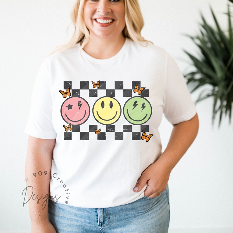 May include: White t-shirt with a checkered pattern and three smiley faces with different expressions. The smiley faces are in pink, yellow, and green. There are orange butterflies around the smiley faces.