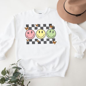 May include: White sweatshirt with a checkered pattern and three smiley faces. The smiley faces are pink, yellow, and green. Each smiley face has a different design. The pink smiley face has a star and a lightning bolt. The yellow smiley face has a simple smile. The green smiley face has two lightning bolts. There are orange butterflies around the smiley faces.