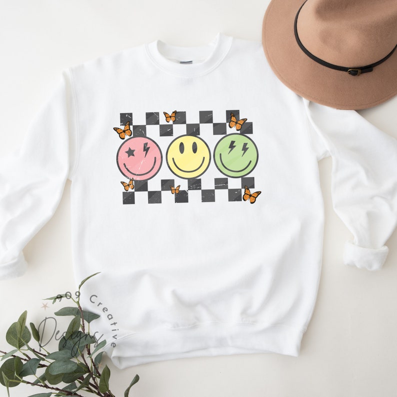 May include: White sweatshirt with a checkered pattern and three smiley faces. The smiley faces are pink, yellow, and green. Each smiley face has a different design. The pink smiley face has a star and a lightning bolt. The yellow smiley face has a simple smile. The green smiley face has two lightning bolts. There are orange butterflies around the smiley faces.