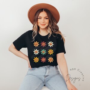 Puede incluir: Una camiseta negra con un patr&oacute;n colorido de caras de calabaza sonrientes dentro de flores. Las flores son en tonos de naranja, amarillo, verde y marr&oacute;n. La camiseta la lleva una persona con cabello casta&ntilde;o y un sombrero marr&oacute;n.
