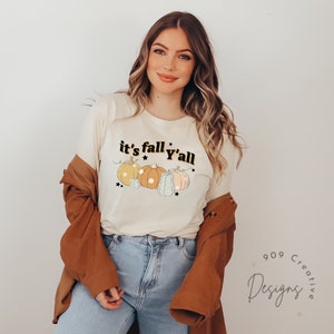 Puede incluir: Una camiseta blanca con el texto "it's fall y'all" en letras negras y doradas. La camiseta presenta una l&iacute;nea de cinco calabazas en varios tonos de naranja, amarillo y azul con contornos blancos. Las calabazas est&aacute;n decoradas con peque&ntilde;as estrellas.