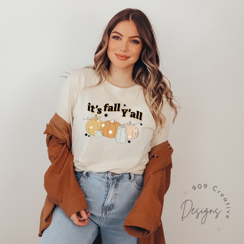 Puede incluir: Una camiseta blanca con el texto "it's fall y'all" en letras negras y doradas. La camiseta presenta una l&iacute;nea de cinco calabazas en varios tonos de naranja, amarillo y azul con contornos blancos. Las calabazas est&aacute;n decoradas con peque&ntilde;as estrellas.