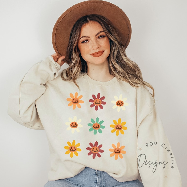 Puede incluir: Una sudadera de color crema con un patr&oacute;n de nueve flores naranjas, amarillas, rojas y verdes con caras de calabaza. El texto "909 Creative Designs" est&aacute; en la esquina inferior derecha de la sudadera.