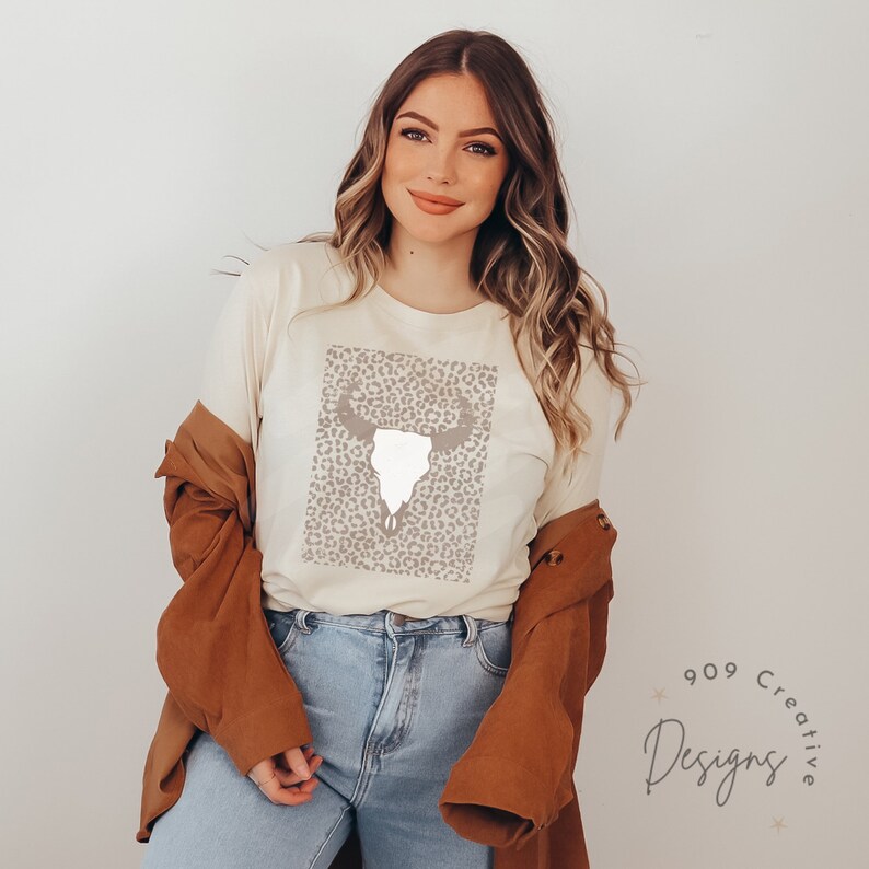 May include: A white t-shirt with a brown and white leopard print design featuring a white skull with antlers. The skull is centered on the print. The t-shirt is being worn by a person.