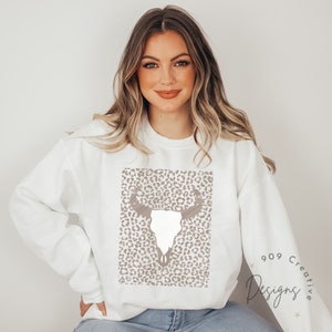 May include: White sweatshirt with a brown and white leopard print design featuring a white skull with horns. The text "909 Creative Designs" is in the bottom right corner of the image.