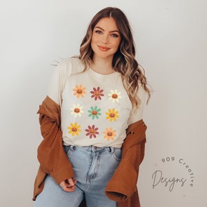 Puede incluir: Una camiseta blanca con un patr&oacute;n de nueve flores de colores. Cada flor tiene una cara de calabaza en el centro.