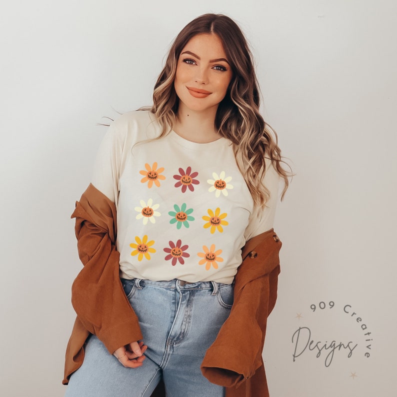 Puede incluir: Una camiseta blanca con un patr&oacute;n de nueve flores de colores. Cada flor tiene una cara de calabaza en el centro.