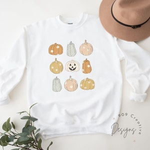 May include: White sweatshirt with a design of nine pumpkins in various colors and patterns. The pumpkins are arranged in a grid pattern. The sweatshirt has a crew neck and long sleeves.