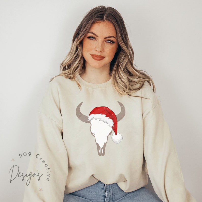 May include: A cream coloured sweatshirt with a grey bull skull wearing a red Santa hat with white trim. The text "909 Creative Designs" is on the bottom left of the sweatshirt.