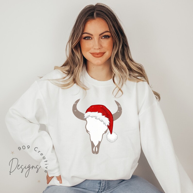 May include: White sweatshirt featuring a brown bull skull wearing a red Santa hat with white trim. The sweatshirt has the text "909 Creative Designs" on the bottom left corner.