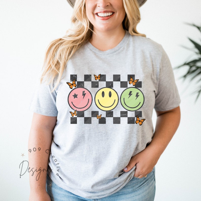 May include: A gray t-shirt with a checkered pattern and three smiley faces. The smiley faces are pink, yellow, and green. Each smiley face has a different design, including a star, a lightning bolt, and two lightning bolts. There are orange butterflies around the smiley faces.