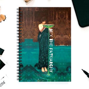 May include: Spiral-bound notebook featuring a teal and brown artistic design with a woman holding a bowl. The text "HEX THE PATRIARCHY" is printed vertically on the right side of the notebook.