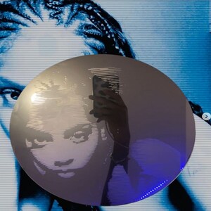 Op de afbeelding: Een ronde zilveren spiegel met een gezicht dat erin wordt weerspiegeld. Het gezicht is gemaakt van lijnen en lijkt een digitale afbeelding te zijn. De spiegel is omringd door een blauw licht.