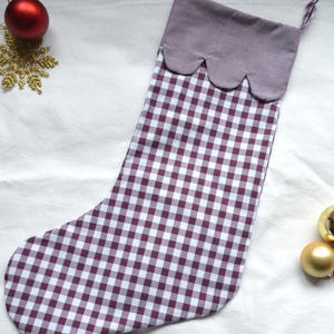 May include: A Christmas stocking featuring a white and dark purple gingham check pattern on the main body. The cuff is a solid dark purple fabric with a scalloped bottom edge and a small matching gingham loop attached to the top corner for hanging.