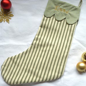 May include: A Christmas stocking featuring a cream and olive green vertical striped body with a scalloped cuff in a green and white gingham pattern. The cuff is embroidered with the name Emma in gold thread and includes a small hanging loop.