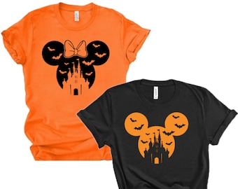 May include: Two t-shirts with a Halloween-themed Mickey Mouse design. The orange shirt features a black silhouette of Mickey Mouse with a bow and bats flying around a castle. The black shirt features a black silhouette of Mickey Mouse with bats flying around a castle in an orange circle.