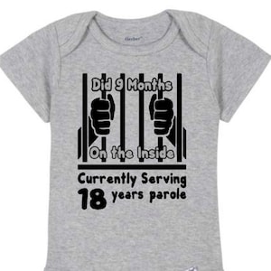 May include: Gray baby onesie with black text that reads "Did 9 Months On the Inside Currently Serving 18 years parole" with a black jail cell graphic.