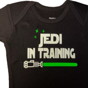 May include: Black baby onesie with white text that reads "Jedi in Training" and a green lightsaber graphic.