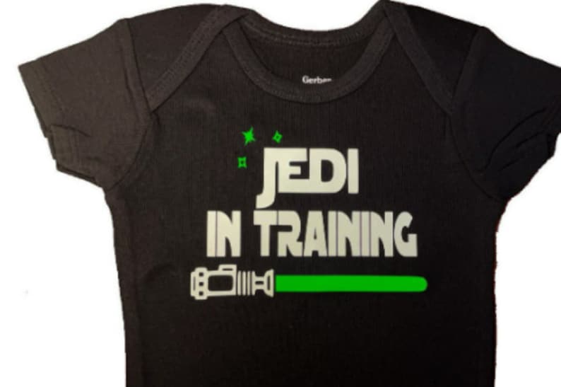May include: Black baby onesie with white text that reads "Jedi in Training" and a green lightsaber graphic.