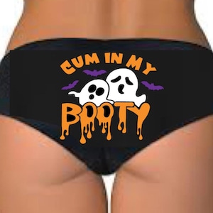 May include: Black underwear with the text "CUM IN MY BOOTY" in orange and white, with two cartoon ghosts and purple bats. The underwear is a cheeky style.