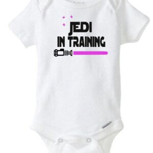May include: White baby onesie with black text that reads "Jedi in Training" and a pink lightsaber graphic.