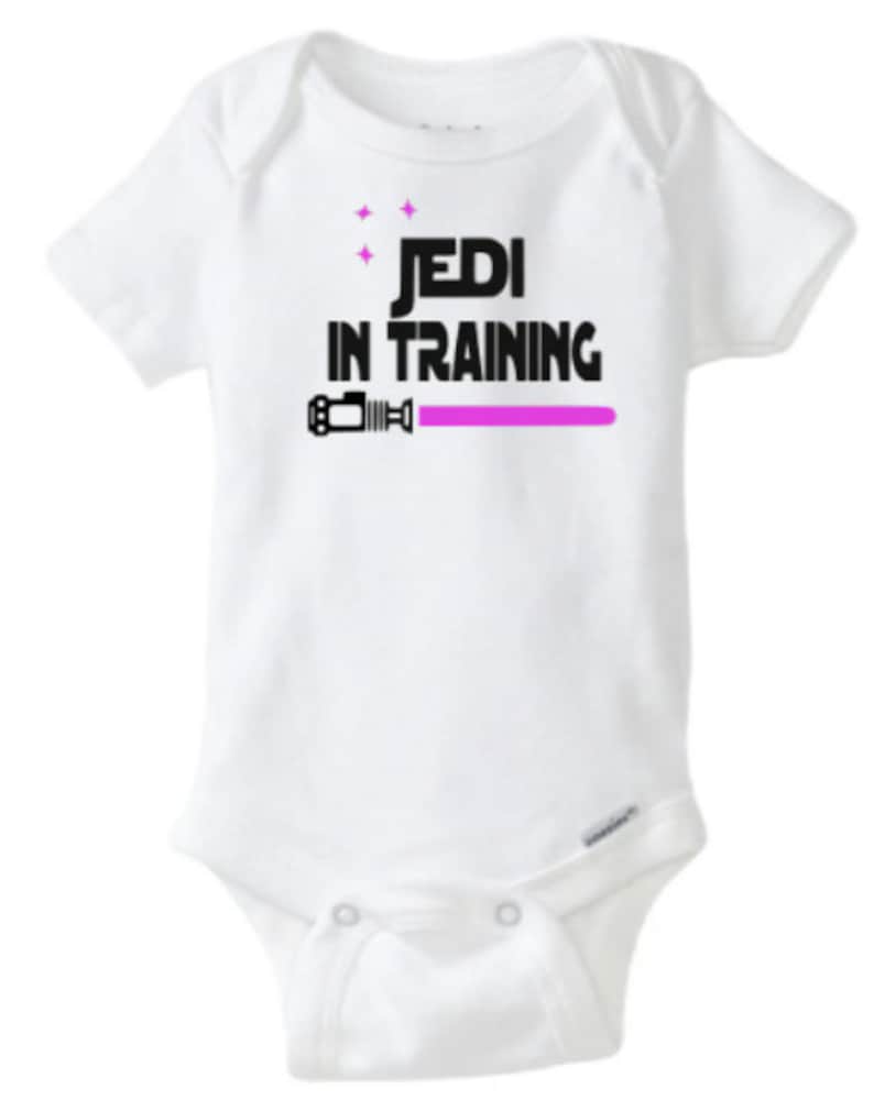 May include: White baby onesie with black text that reads "Jedi in Training" and a pink lightsaber graphic.