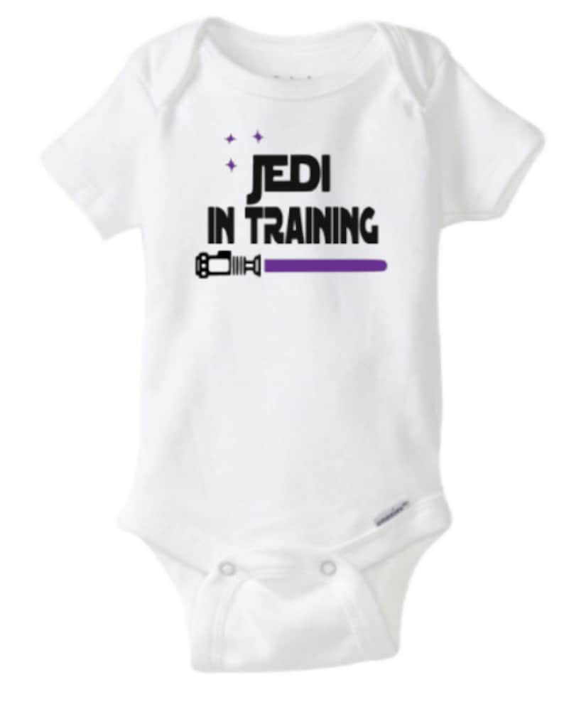 May include: White baby onesie with black text that reads "Jedi in Training" and a purple lightsaber graphic.