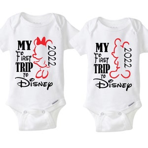 May include: Two white baby onesies with the text "MY FIRST TRIP to Disney 2022". One onesie features a red Minnie Mouse silhouette, and the other features a red Mickey Mouse silhouette. The text is in black and red.