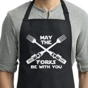 May include: Black apron with white text that reads "May the Forks Be With You". Two crossed forks are depicted with lightsaber blades.