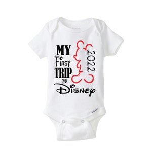 May include: White baby onesie with short sleeves. The onesie has the text "MY FIRST TRIP TO Disney 2022" in black and red. A red outline of Mickey Mouse is on the right side of the text.