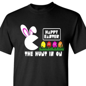 May include: Black t-shirt with a white Pac-Man character with pink bunny ears eating colorful Easter eggs. The text "Happy Easter" and "The Hunt Is On" is printed on the shirt.