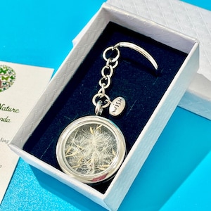 May include: A silver keychain with a round glass locket containing dried dandelion seeds. The locket is attached to a silver chain with a small silver tag that says "Wish". The keychain is in a white box with a black interior.