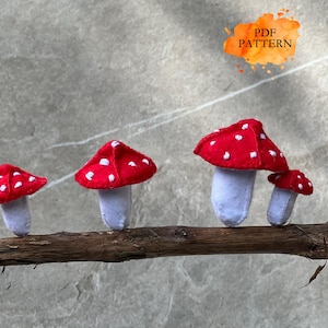 May include: Four felt mushroom decorations with red caps and white spots, perched on a brown branch. The mushrooms have white stems. The image includes the text "PDF PATTERN" in an orange watercolor-style shape.