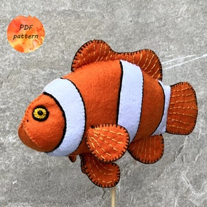 May include: A handmade felt clownfish in orange, white, and gold. The fish has a white stripe around its body and gold stitching details. The image includes a small circular graphic with the text "PDF pattern".