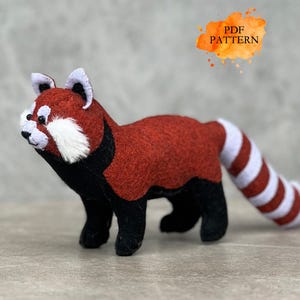 Può includere: Figurina di panda rosso in feltro fatta a mano con petto e viso bianchi, gambe e dettagli neri e coda a strisce. L'immagine include una sovrapposizione di testo in stile acquerello che dice "PDF PATTERN".