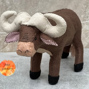 Op de afbeelding: Een bruin en wit gevuld knuffeldier van vilt in de vorm van een buffel met zwarte hoeven. De buffel heeft grote, witte hoorns en een roze neus. De tekst "PDF pattern" staat op een aquarelcirkel in de linkerbenedenhoek van de afbeelding.