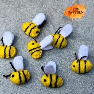 Puede incluir: Un conjunto de abejas de fieltro hechas a mano en amarillo y negro, con alas blancas y antenas negras. Las abejas están dispuestas sobre una superficie gris. La imagen también incluye un gráfico de acuarela naranja y amarillo con el texto "PDF PATTERN".