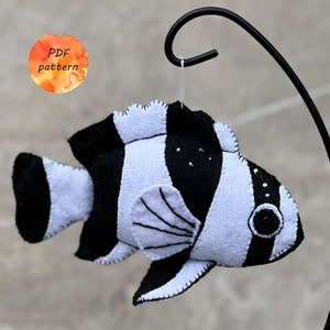 Könnte beinhalten: Handgefertigter Fischschmuck aus Filz in schwarz-weißen Streifen, mit einer genähten Kontur. Der Fisch hängt an einem schwarzen Metallständer. Das Bild enthält einen Aquarellkreis mit dem Text "PDF pattern".
