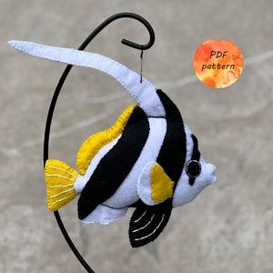 May include: A handmade felt fish ornament with a black, white, and yellow color scheme. The fish has a long, white dorsal fin and a yellow tail fin. The ornament is hanging from a black metal stand. The image includes a small orange circle with the text "PDF pattern".