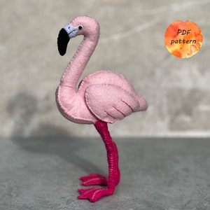 May include: A handmade pink felt flamingo with a black beak and white accents. The flamingo has a long, curved neck and stands on bright pink legs and feet. A small orange circle with the text "PDF pattern" is in the background.