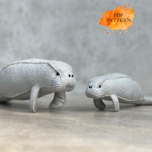 Puede incluir: Dos peluches de manatíes de fieltro gris. El manatí más grande está delante, con el más pequeño detrás. Los manatíes tienen pequeños ojos negros y detalles cosidos. La imagen incluye el texto "PDF PATTERN" en un salpicadura de acuarela naranja.