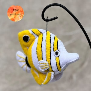 May include: A felt fish decoration in yellow, white, and black, hanging from a black metal hook. The fish has yellow stripes and a detailed eye. A watercolour circle with the text "PDF pattern" is in the background.