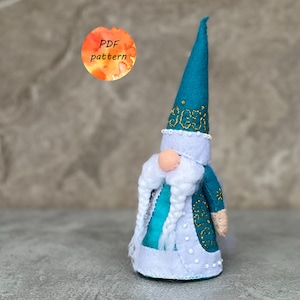 Op de afbeelding: Een handgemaakte kabouterpop met een teal puntmuts en bijpassende tuniek, versierd met gouden borduursel. De kabouter heeft een lange witte baard, een perzikkleurige neus en een wit afgewerkte jas. De afbeelding bevat een aquarel cirkel met de tekst "PDF pattern".