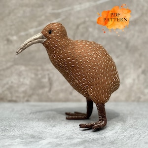 Può includere: Una statuetta di uccello kiwi in tessuto marrone con dettagli cuciti bianchi sul corpo, un lungo becco curvo di colore chiaro e zampe marrone scuro. Un grafico in stile acquerello arancione nell'angolo in alto a destra mostra il testo PDF PATTERN.