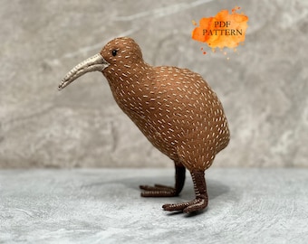 Può includere: Una statuetta di uccello kiwi in tessuto marrone con dettagli cuciti bianchi sul corpo, un lungo becco curvo di colore chiaro e zampe marrone scuro. Un grafico in stile acquerello arancione nell'angolo in alto a destra mostra il testo PDF PATTERN.