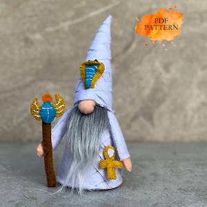 Op de afbeelding: Een lichtpaarse viltpop in de vorm van een kabouter met een hoge puntmuts en een goudkleurig en blauw cobra-embleem. De kabouter heeft een lange grijze baard, houdt een bruine staf met een blauwe scarabee vast en draagt een gouden ankh-symbool. Tekst PDF PATTERN.