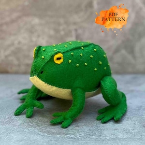 Könnte beinhalten: Eine handgefertigte grüne Filzfrosch mit gelben Augen und einem hellgelben Bauch. Der Frosch hat kleine, dekorative gelbe Stiche auf dem Rücken. Das Bild enthält eine orangefarbene und gelbe Textblase im Aquarellstil mit der Aufschrift "PDF PATTERN".