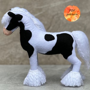 Puede incluir: Un caballo de peluche blanco y negro con manchas, con una larga melena y cola blancas. El caballo tiene patas blancas con plumas y una nariz rosa. La imagen incluye el texto "PDF pattern".