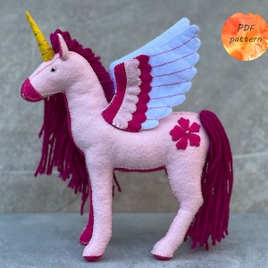 Peut inclure: Une licorne en feutre rose avec une corne dorée, des ailes roses et bleues et une fleur rose sur le côté. La licorne a une longue crinière et une queue roses.